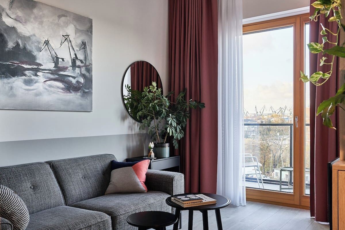 A modern living room with a grey sofa, a round mirror, a potted plant, and a view through a large window.
