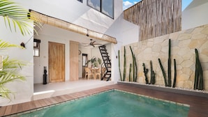 Un'area piscina moderna all'aperto con un solarium in legno, un muro in pietra con cactus e una scala che conduce a un livello superiore.
