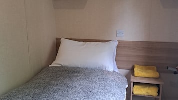 A single bed with a grey textured blanket, a white pillow, and a wooden headboard. A small wooden nightstand with a yellow towel is to the right.
