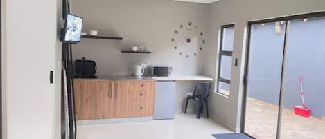 Cocina compacta con microondas, hervidor y refrigerador pequeño. Hay un armario de madera y una silla cerca de la ventana.