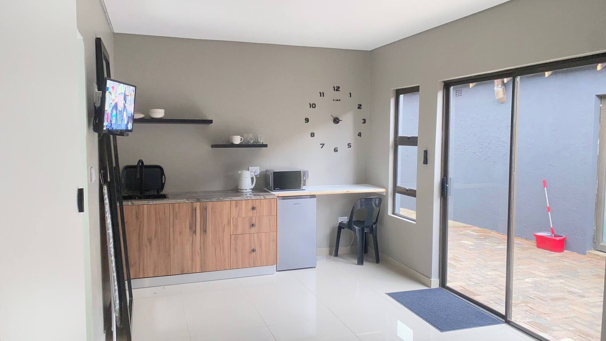 A compact kitchen with a microwave, toaster, and coffee maker on the counter, a small refrigerator, and a wall clock.