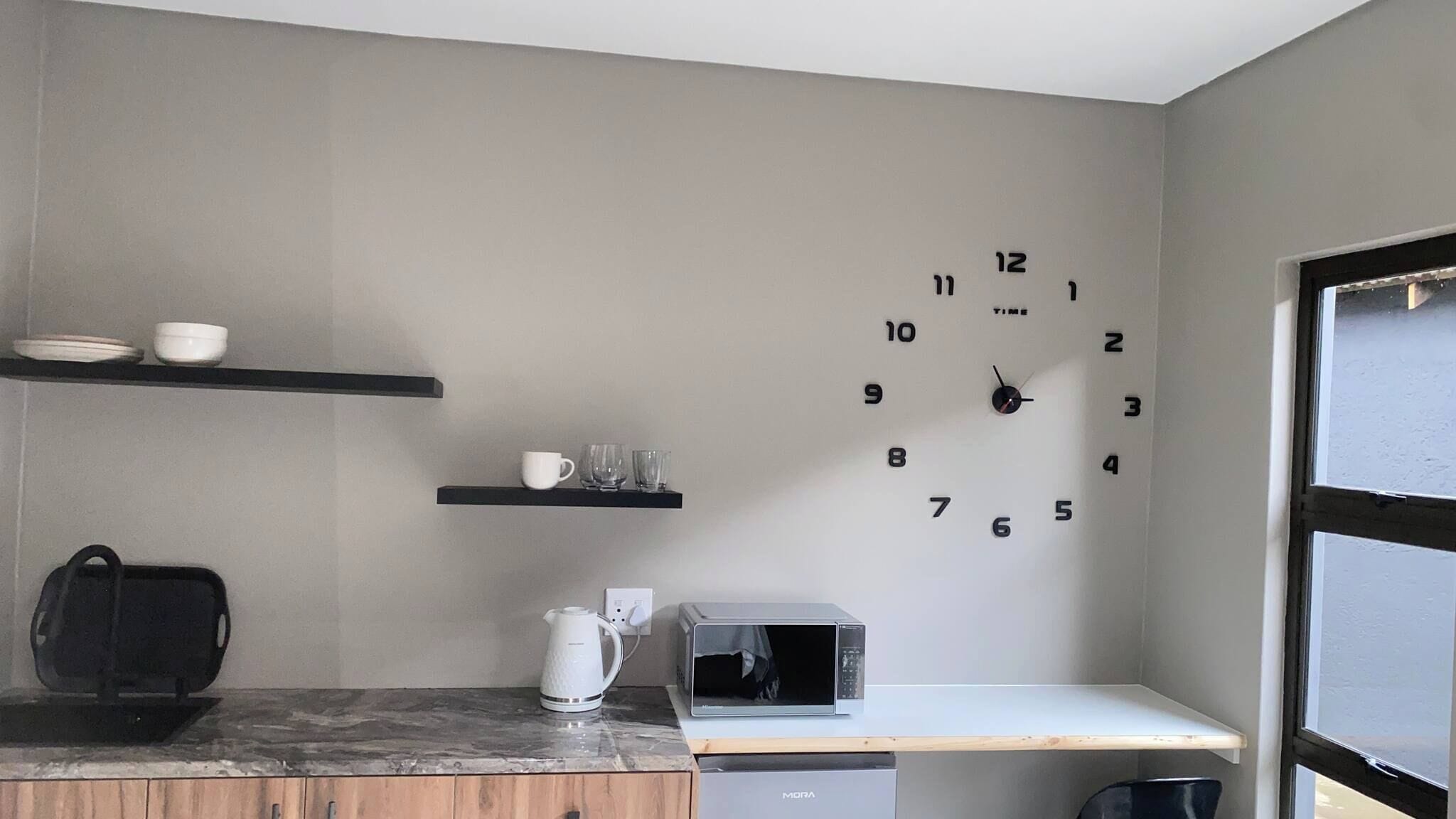 Una cocina con un reloj de pared, estantes con vajilla, un hervidor y un microondas.