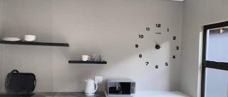 Uma cozinha com relógio na parede, prateleiras com louças, chaleira e micro-ondas.