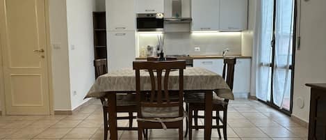 Cocina compacta con armarios blancos, una mesa de comedor con sillas y una puerta corrediza de vidrio a la derecha.