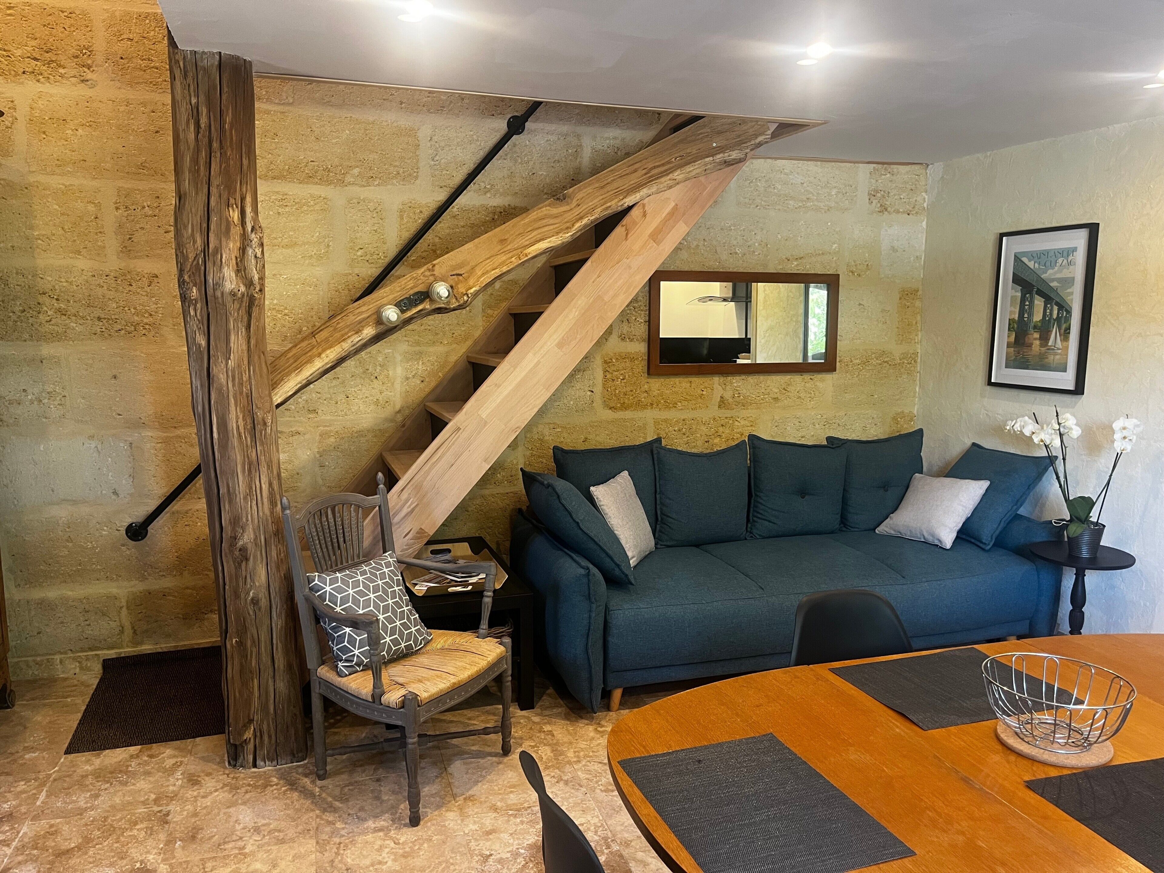 Una habitación con escalera de madera, un sofá azul, una mesa de comedor con individuales y una silla con cojín estampado.