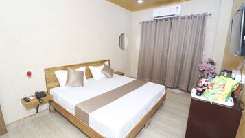 Une chambre d’hôtel comprenant un lit, une table de chevet avec un téléphone, un miroir, un appareil de climatisation, des rideaux, une télévision et une petite table avec des collations et des boissons.