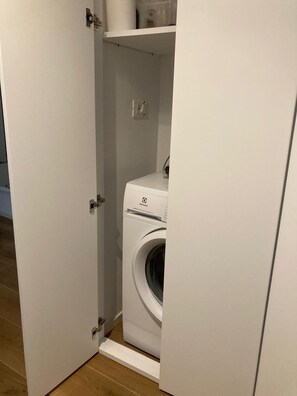 A compact, white washing machine in a small, enclosed laundry room with a door and a shelf above it. - "42 square meters"A STYLISH BRAND-NEW FLAT IN THE CITY CENTER IDEAL FOR FAMILIES (BARI)