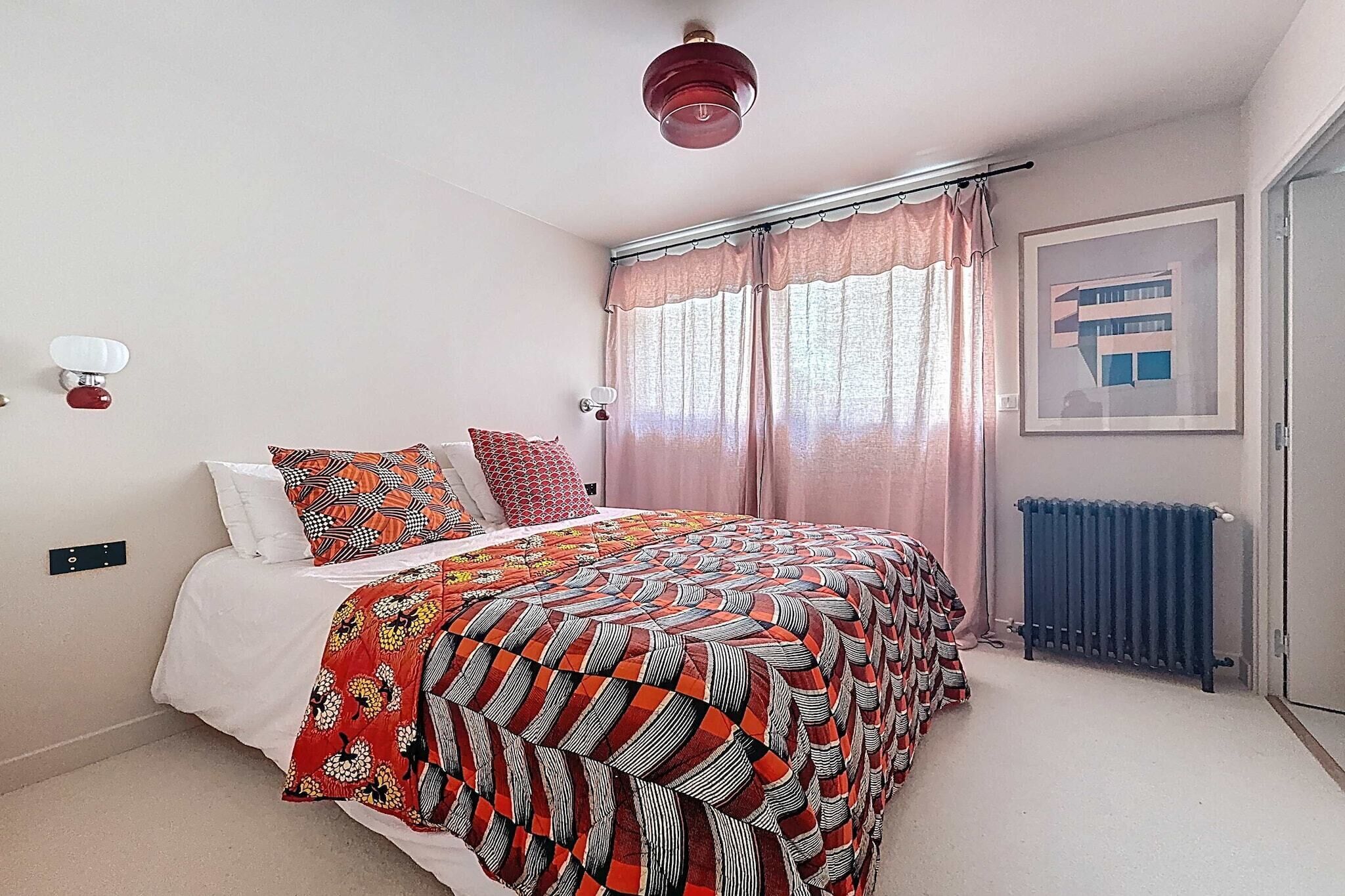 A bedroom with a bed, a radiator, a window with curtains, and a wall-mounted light fixture.