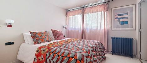 A bedroom with a bed, a radiator, a window with curtains, and a wall-mounted light fixture.