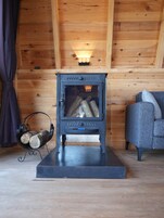 Una estufa de leña con leños, un portaleños metálico y un sofá gris en una habitación de madera.