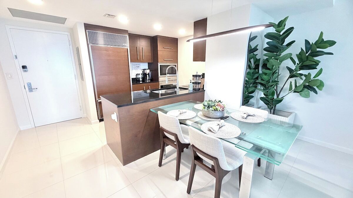 A modern kitchen with a central island, a dining area with a glass table, and a large potted plant.