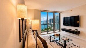A modern living room with a sofa, floor lamps, a glass coffee table, a flat-screen TV, and a large window offering a city view. - Skyline Views 41st Fl Luxury 1BD @ ICON Brickell (Miami)