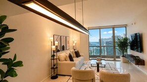 Ein modernes Wohnzimmer mit Sofa, Sesseln, Couchtisch und Fernseher. An den Wänden befinden sich Pflanzen und Kunstwerke.
