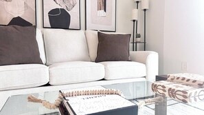 Ein modernes Wohnzimmer mit einem weißen Sofa, braunen Kissen, einem Glastisch und gerahmten Kunstwerken an der Wand.