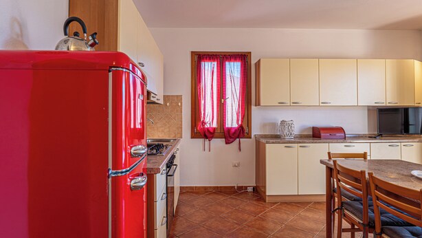 A compact kitchen with a red refrigerator, a stove, and a microwave. There is a window with red curtains, a sink, and a dining area with a table and chairs. - Charming 3-bedroom Apartment in Santa Teresa Gallura (Santa Teresa di Gallura)