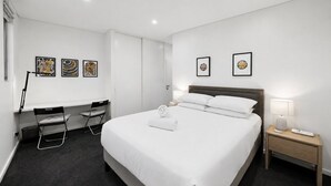 A hotel room with a bed, a desk, two chairs, a nightstand, and artwork on the walls. - Designer 1-Bed Apartment with Balcony (Rozelle)
