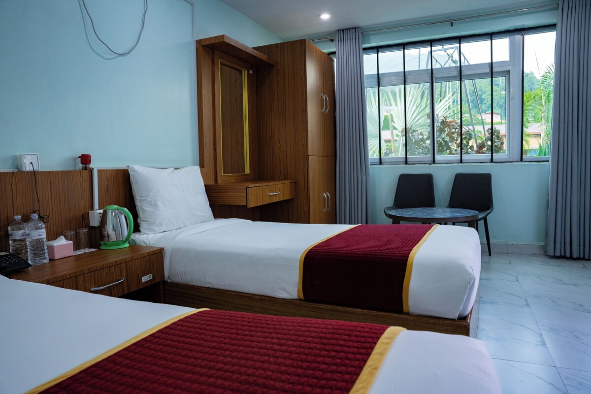 Een hotelkamer met twee bedden, een houten garderobe, een tafeltje met stoelen, een raam met gordijnen en uitzicht op groen.