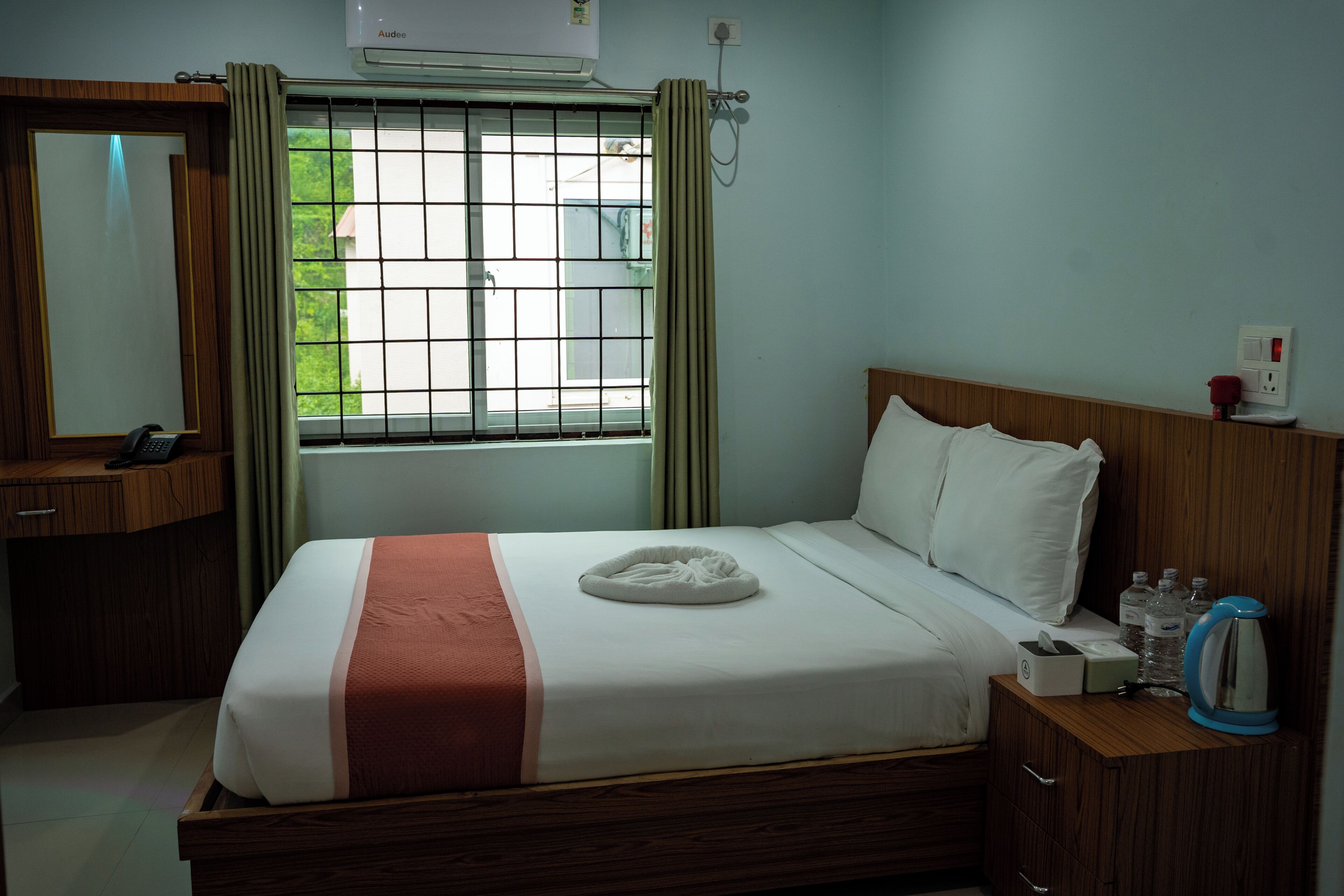 A hotel room with a bed, a bedside table with a kettle, a mirror, and a window with curtains.