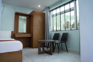 A hotel room with a bed, a wooden wardrobe, a small table with two chairs, and a window with a view of greenery.