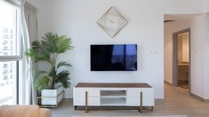 A modern living room with a flat-screen TV mounted on the wall, a white entertainment unit, a large potted plant, and a view of a cityscape through a window. - Bloomfields 3BR plus maid in Waters Edge (Abu Dhabi)