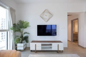 A modern living room with a flat-screen TV mounted on the wall, a white entertainment unit, a large potted plant, and a view of a cityscape through a window. - Bloomfields 3BR plus maid in Waters Edge (Abu Dhabi)