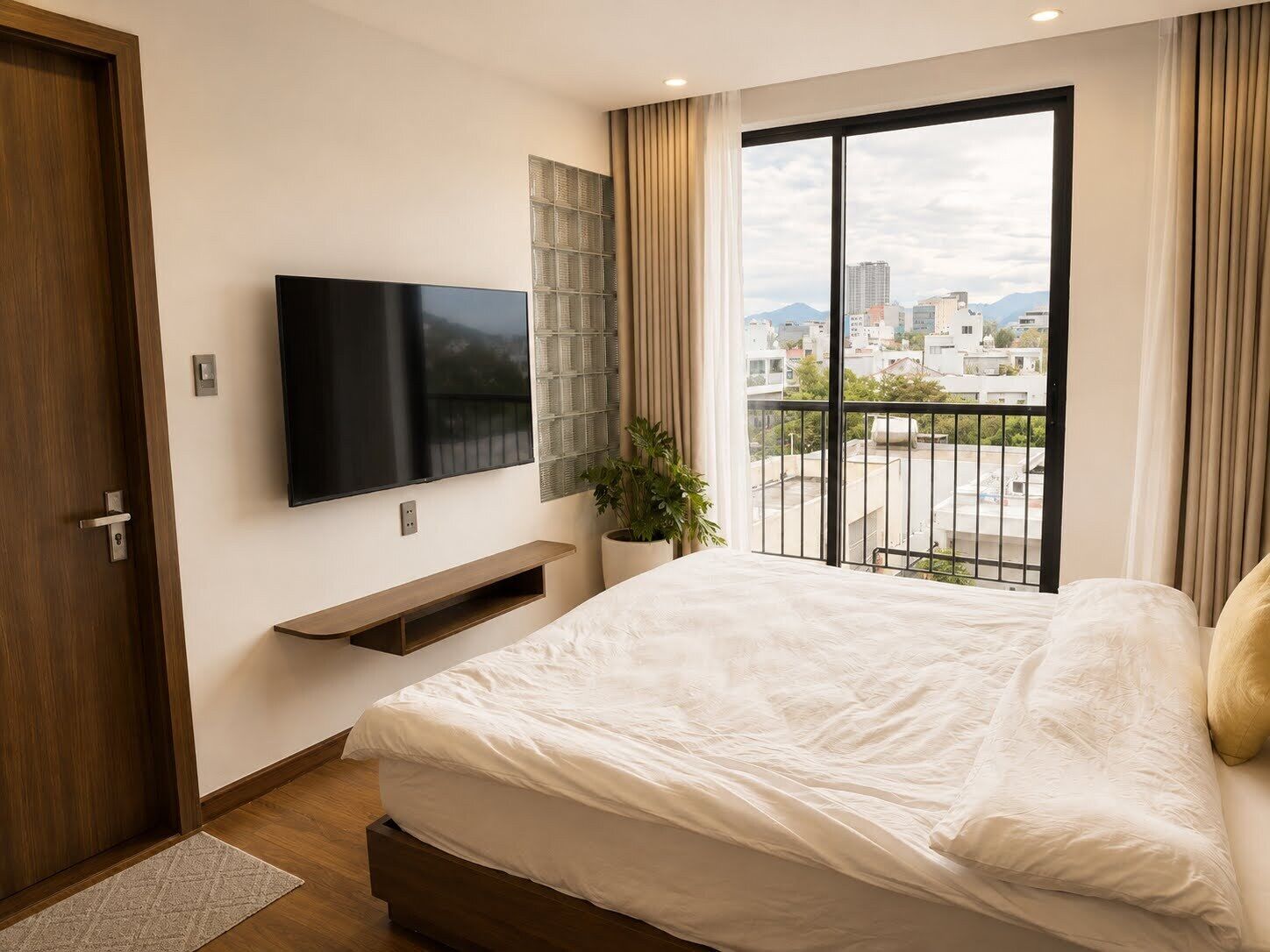 Un dormitorio con cama, televisor montado en la pared, ventana con vista a edificios y puerta.