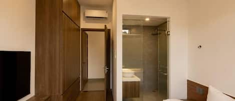 A modern hotel room with a bed, a TV on a wooden shelf, a wardrobe, and a glass-enclosed shower area.