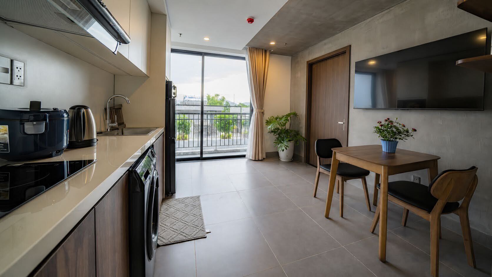 A modern kitchen with a built-in oven, a coffee machine, and a washing machine. There is a dining area with a table and chairs, and a large window offering a view of the outdoors.