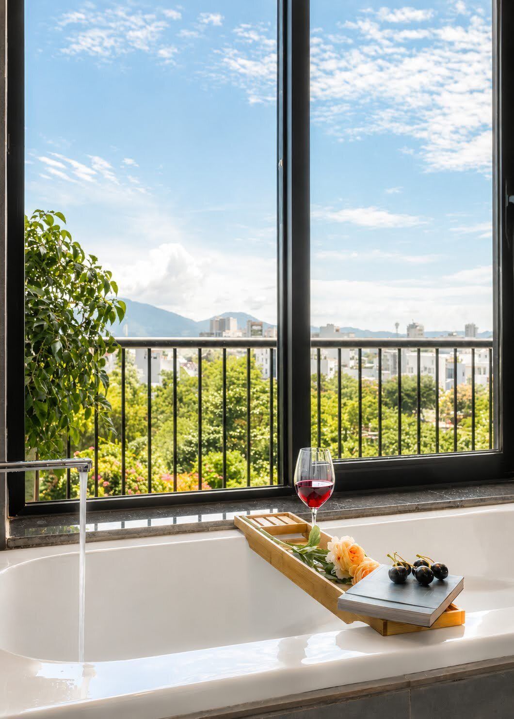 A bathtub with a tray of flowers and a glass of red wine, overlooking a balcony with greenery and a cityscape.