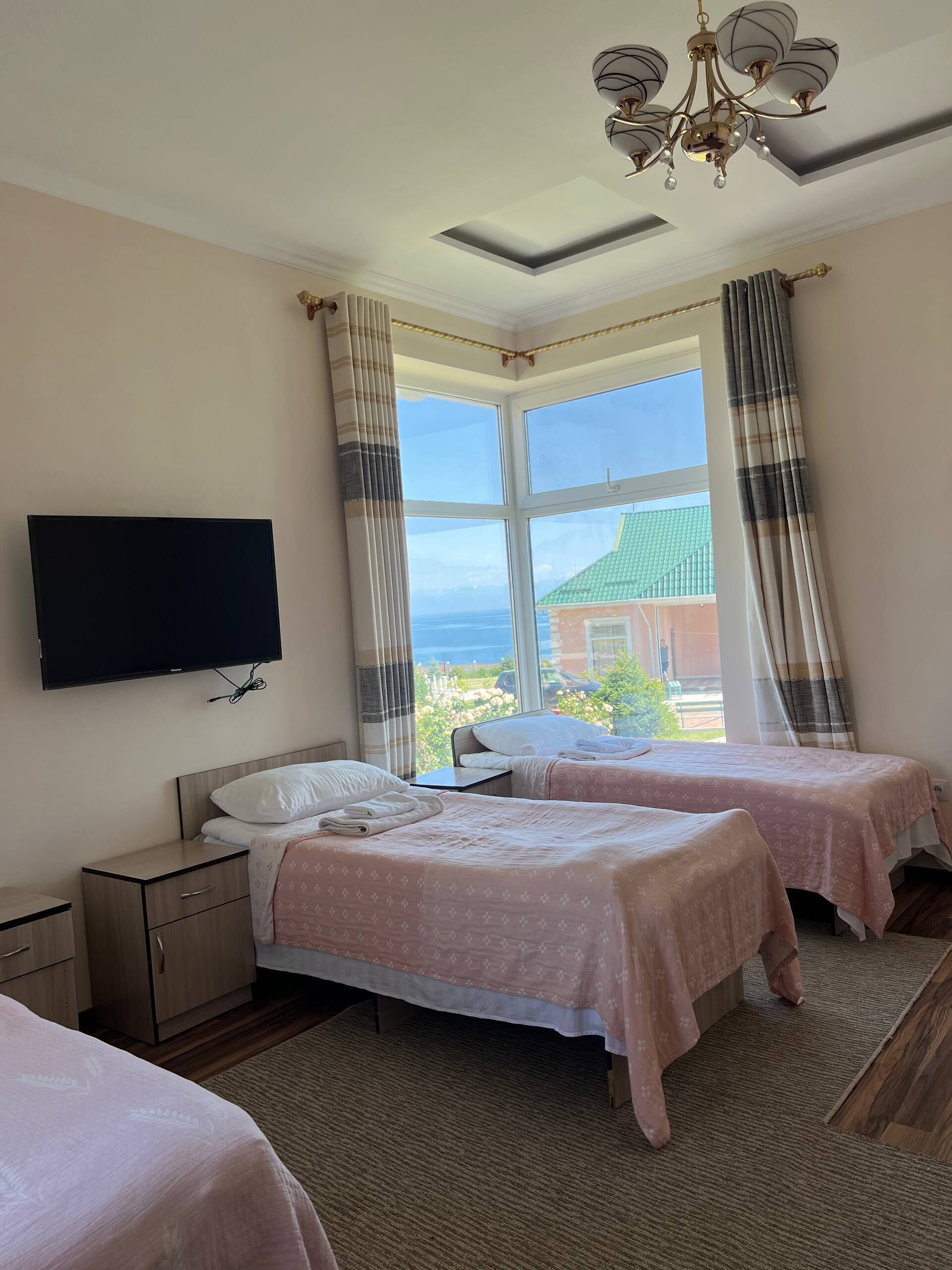 A hotel room with two beds, a TV, a large window with curtains, and a chandelier.