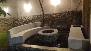 Un espacio de estar al aire libre con forma circular, que incluye un hogar de piedra rodeado por un bajo muro de piedra y un banco curvo blanco.