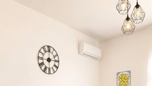 Een woonkamer met een blauwe bank, een wandklok, een airconditioning en een tafel met een decoratief object, plus een plant.