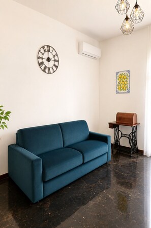 A living room with a blue sofa, a wall clock, an air conditioning unit, a table with a decorative item, and a plant. - Charming Retreat near Historic Landmarks (Modica)