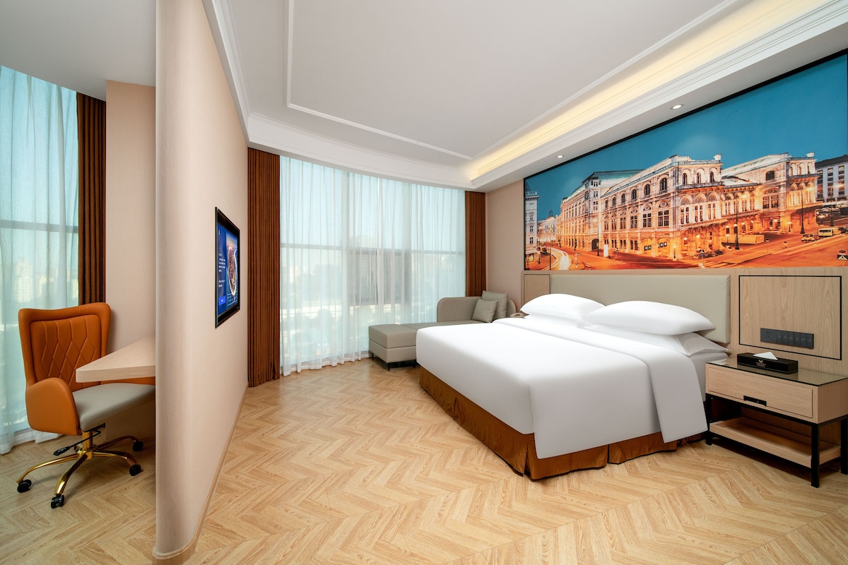 Ein Hotelzimmer mit einem großen Bett, einem Schreibtisch, einem Sessel und einem großen Fenster mit Vorhängen.