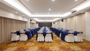 Un salón de conferencias con filas de mesas y sillas, piso alfombrado y una pantalla de proyector al frente.