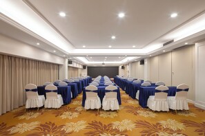 Un salón de conferencias con filas de mesas y sillas, piso alfombrado y una pantalla de proyector al frente.