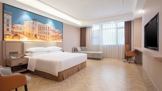 Habitación de hotel con una cama grande, un escritorio, una silla, un televisor y un ventanal con cortinas.