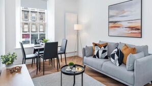 Uma sala de estar moderna com um sofá cinzento, uma mesa de jantar com cadeiras, um abajur e uma janela grande com vista para edifícios.