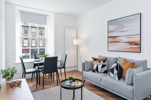 Ein modernes Wohnzimmer mit einem grauen Sofa, einem Esstisch mit Stühlen, einer Stehlampe und einem großen Fenster mit Blick auf Gebäude.