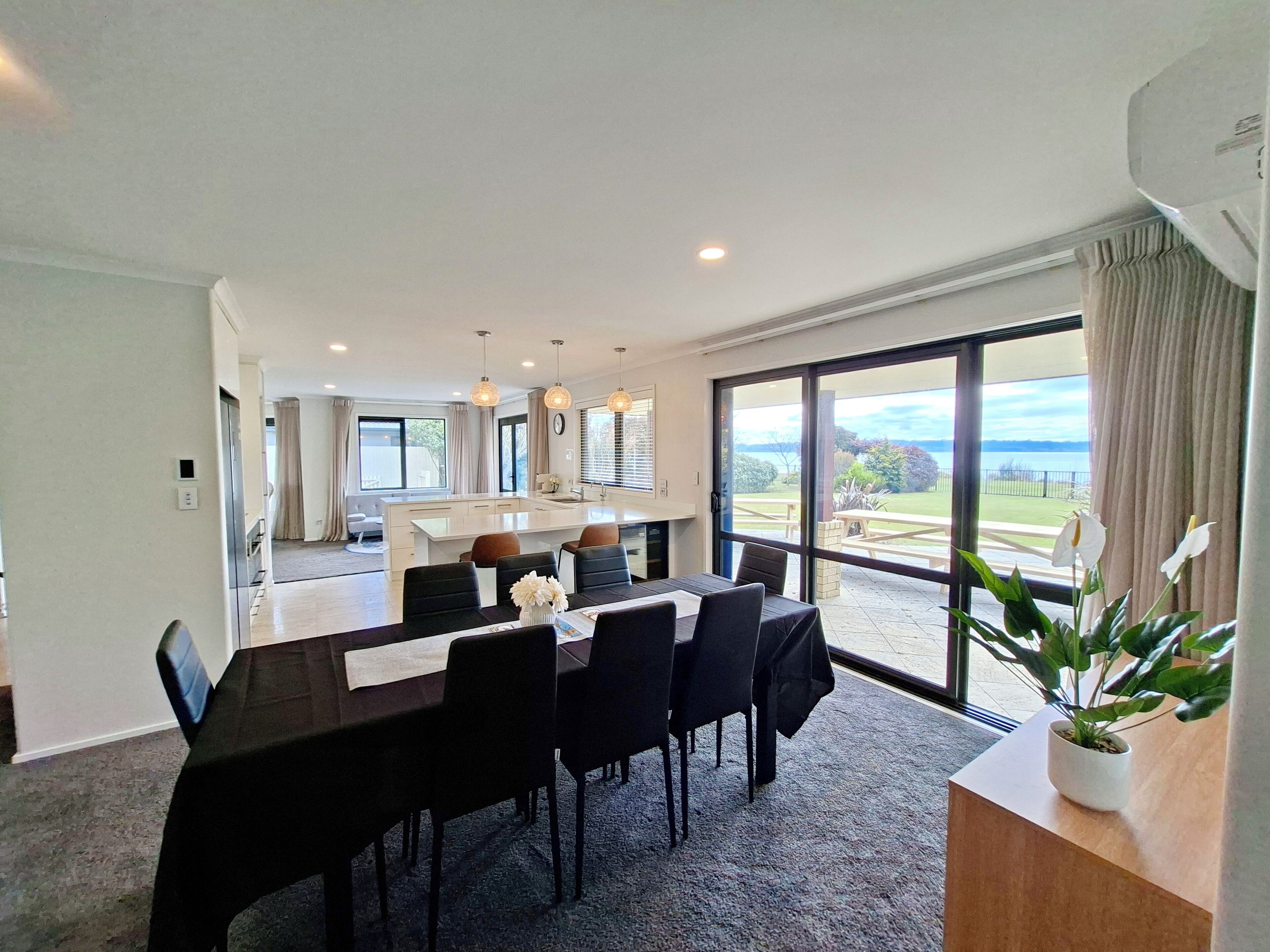 A dining area with a large table and chairs, a kitchen in the background, and a view of the outdoors through large windows.