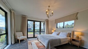 Ein Schlafzimmer mit einem großen Bett, zwei Nachttischen, einer Chandelierleuchtre, Blick durch das Fenster und einem Sessel.