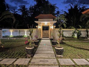 Una entrada bien iluminada con un camino que lleva a un área techada, rodeada de plantas en macetas y palmeras.
