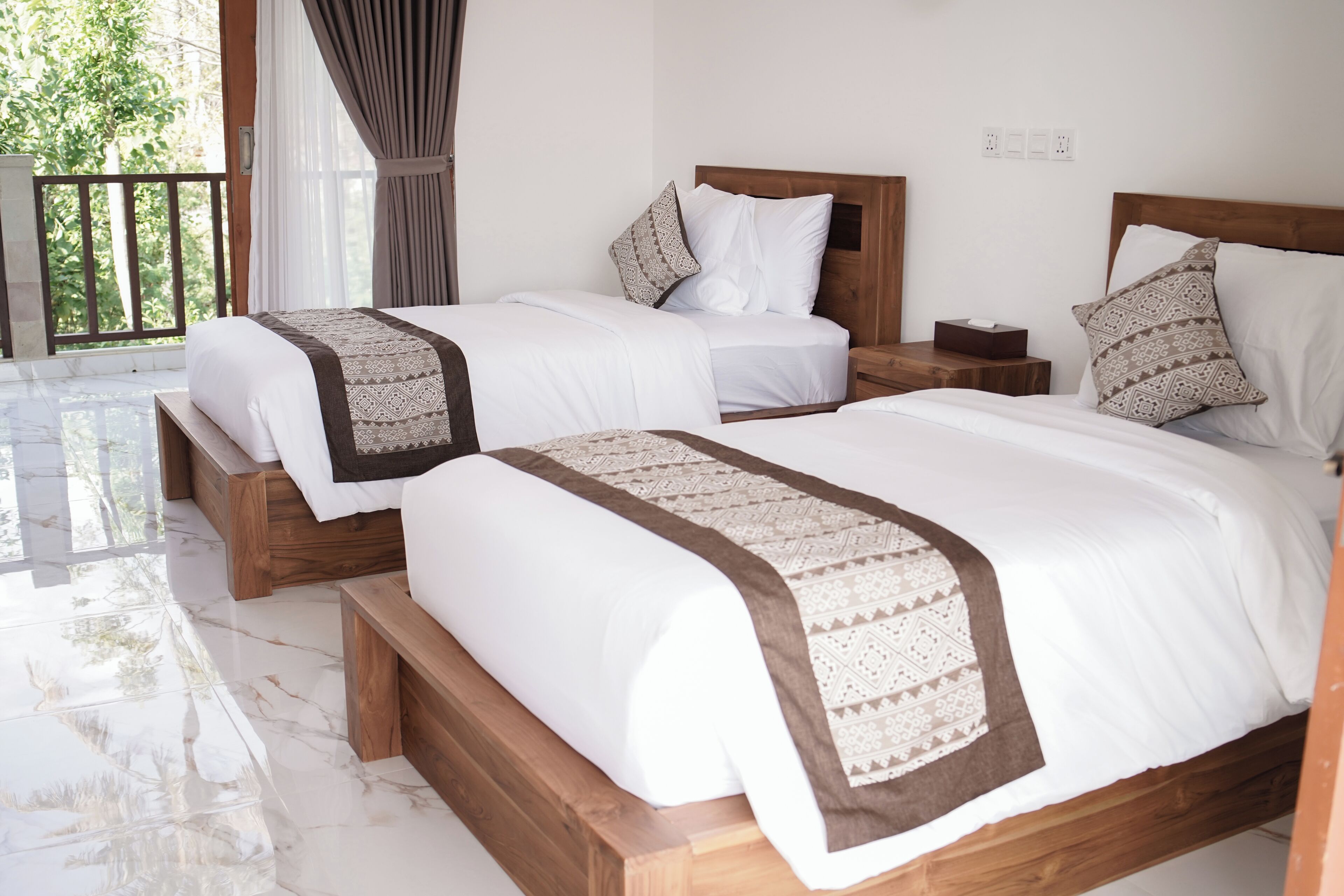 A hotel room with two beds, wooden headboards, white bedding, patterned throw blankets, and a balcony with a view of greenery.
