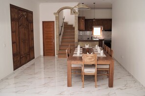 A dining area with a marble floor, wooden table, and chairs. There is a staircase leading to a kitchen with wooden cabinets and a window.