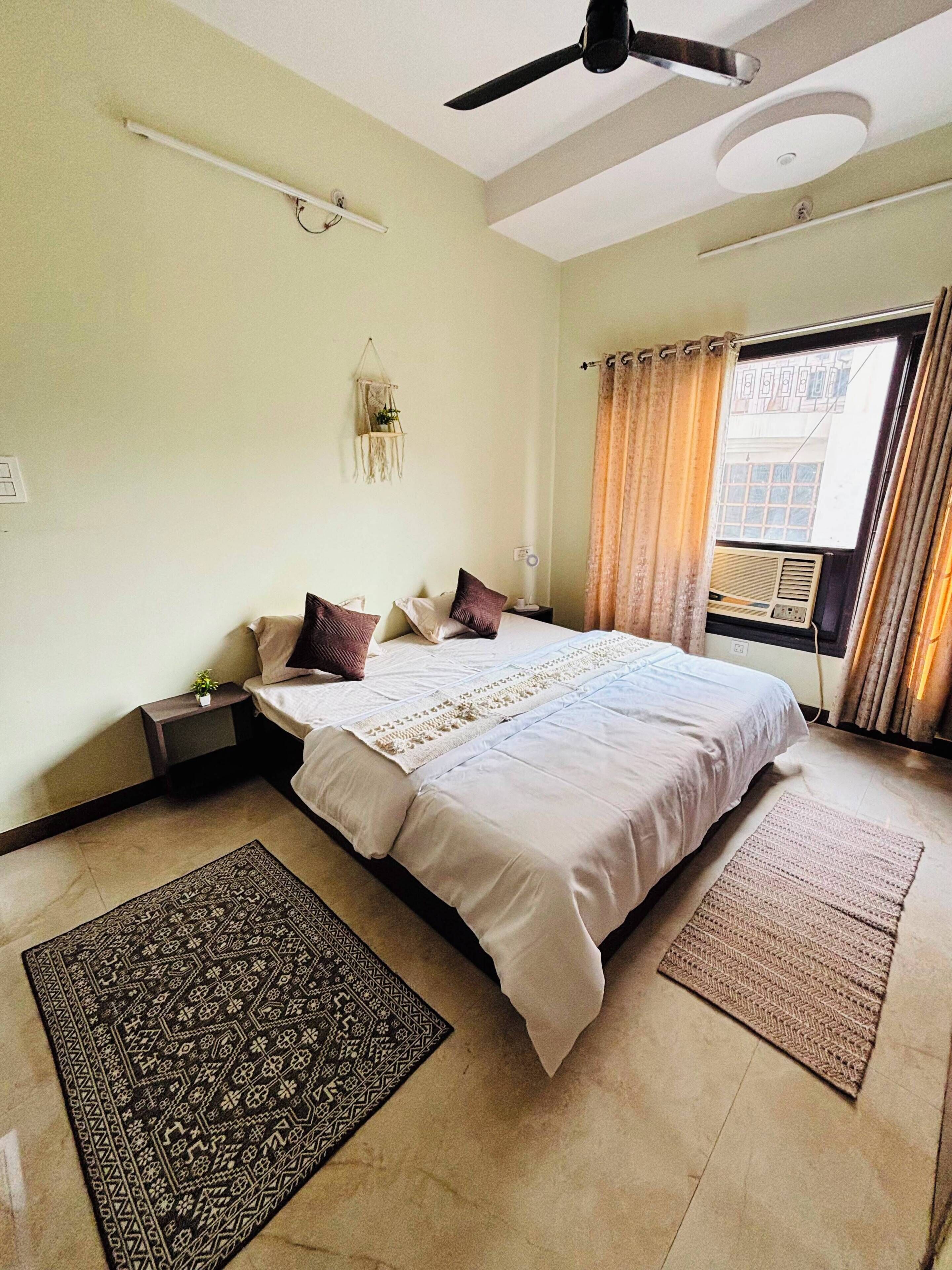 A bedroom with a bed, a ceiling fan, a window with curtains, a wall-mounted shelf, and a small plant.