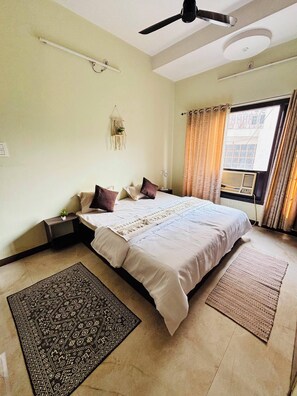 A bedroom with a bed, a ceiling fan, a window with curtains, a wall-mounted shelf, and a small plant.