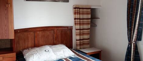 A bedroom with a wooden headboard, a bed with a striped bedspread, a bedside table, a hanging light fixture, a wall-mounted shelf, and a curtain.