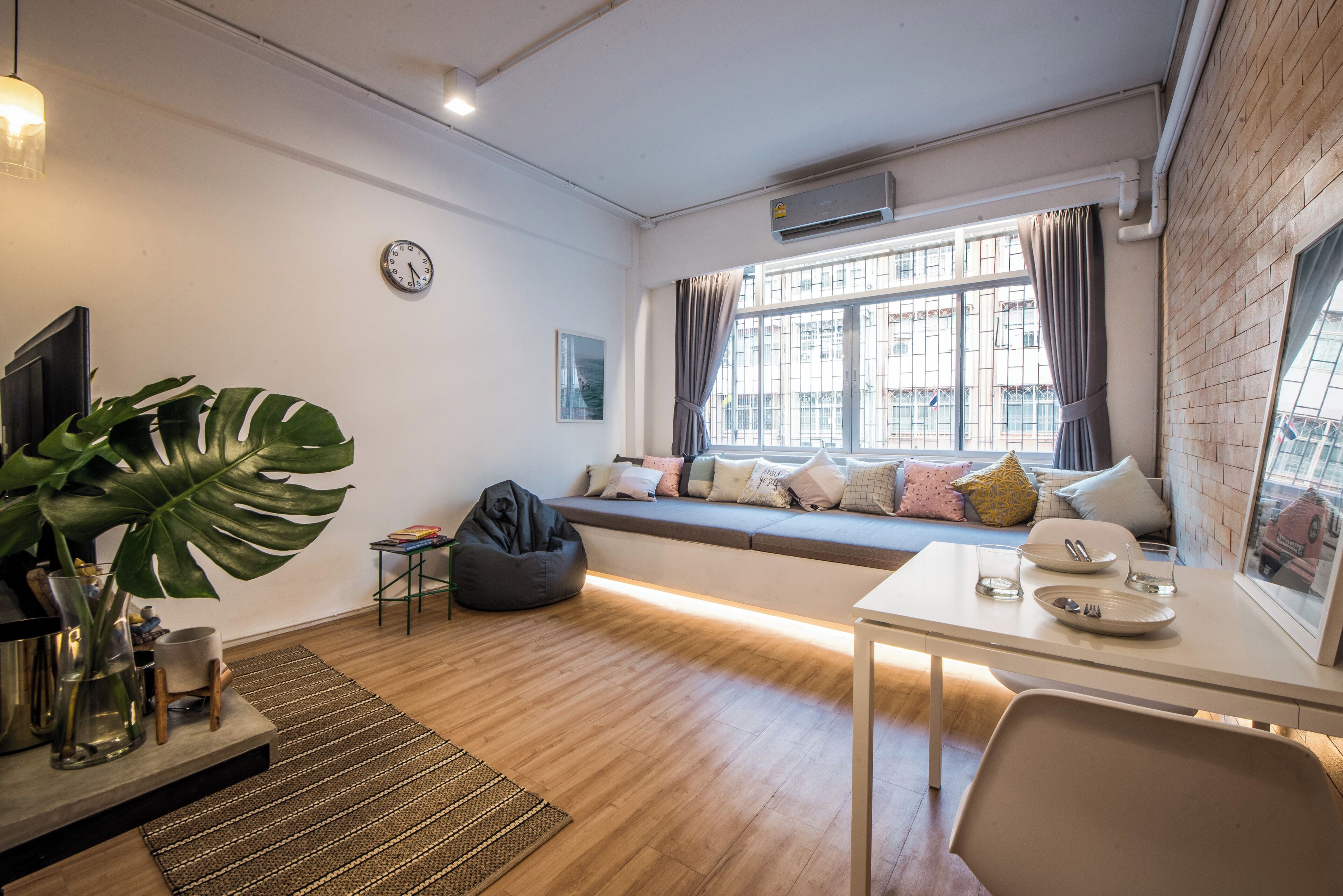 A modern living room with a sofa, a dining table, a large plant, and a view of the outside through a window.
