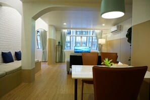 Habitación de hotel con comedor, sofá con cojines, mesa con sillas y un ventanal con cortinas.