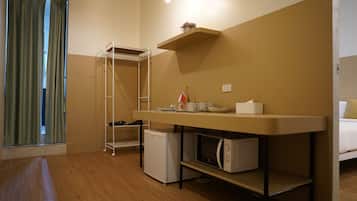 A compact hotel room with a kitchenette, a small refrigerator, a microwave, a shelf with a vase, and a window with curtains.